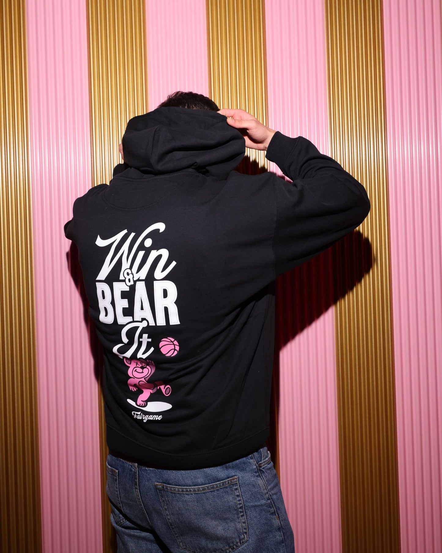 Person wearing a black hoodie with text and graphics on the back against a striped pink and gold wall.