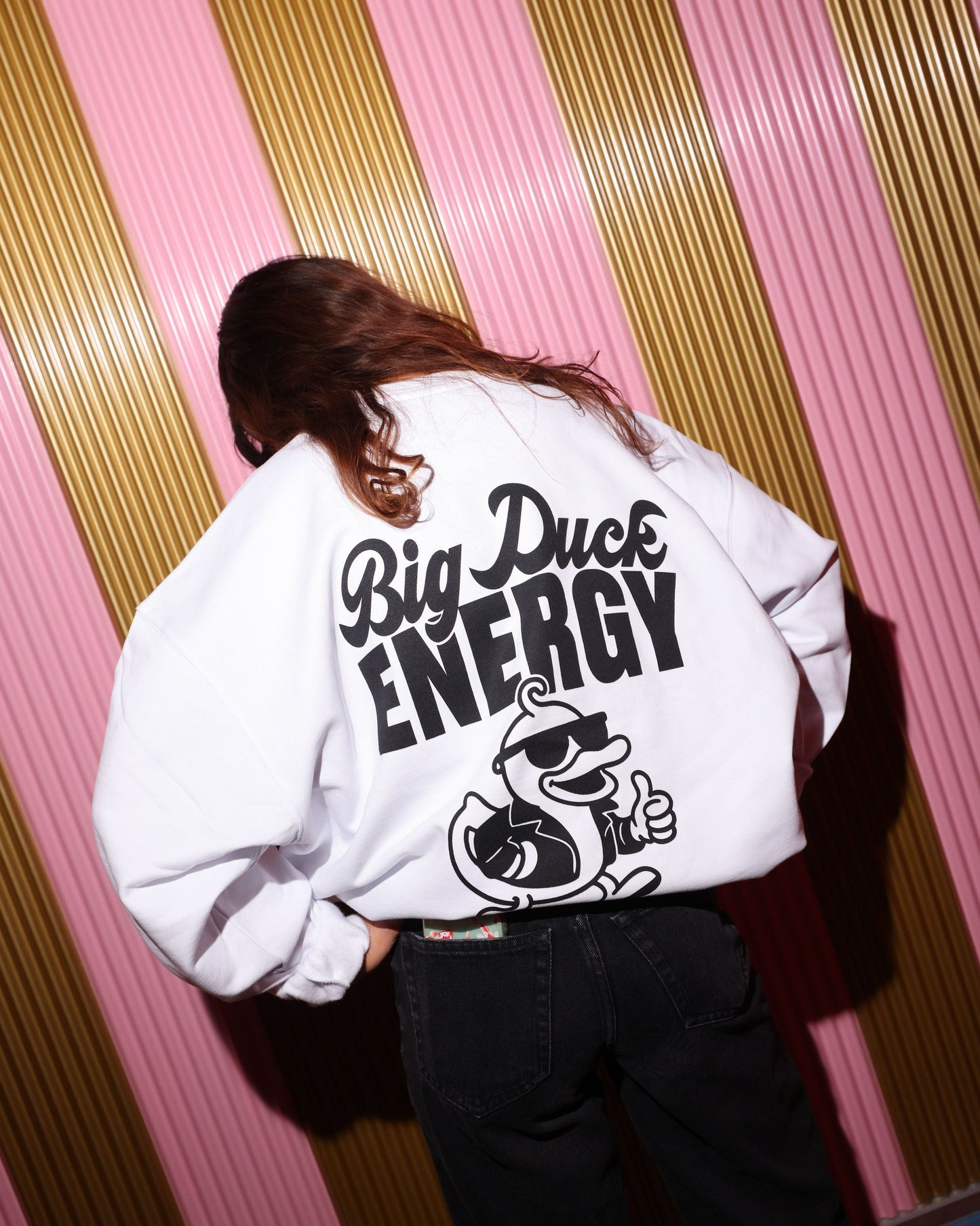 Person wearing a white sweatshirt with 'Big Duck Energy' text and logo against a striped pink and gold wall.