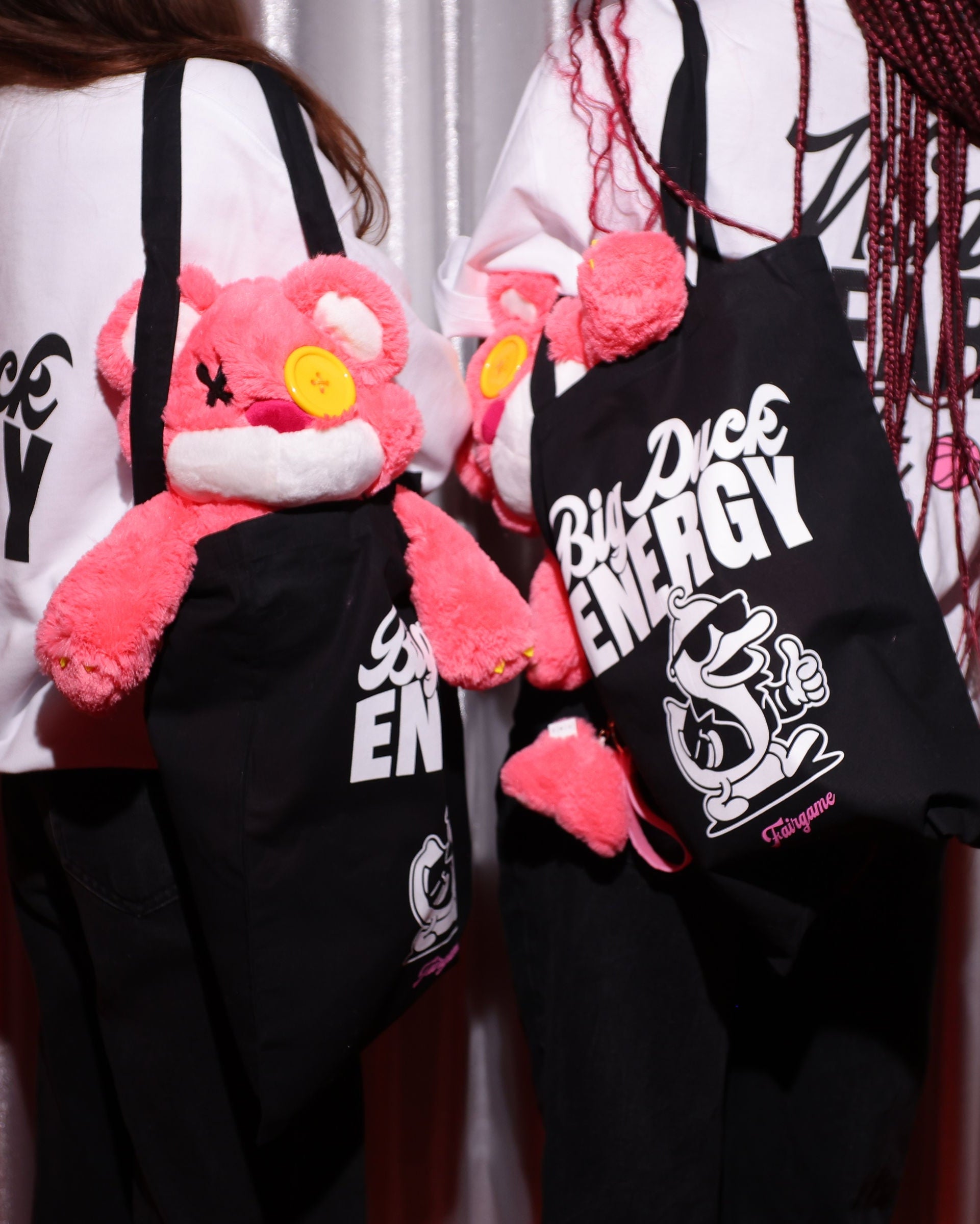 Two people holding black tote bags with pink plush toys against a curtain background